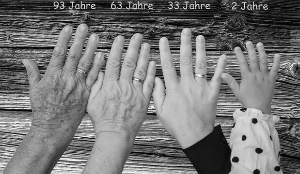 Schwarz-Wei&szlig;aufnahme von vier H&auml;nde im Alter von jeweils 93 Jahren, 63 Jahren, 33 Jahren und 2 Jahren vor einem Hintergrund mit Holzstruktur und Altersangaben in Textform