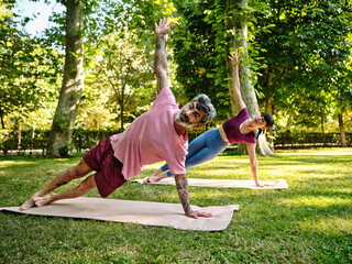 Concentrated couple doing yoga on mats together in Phalakasana with raised arm on sunny day in park