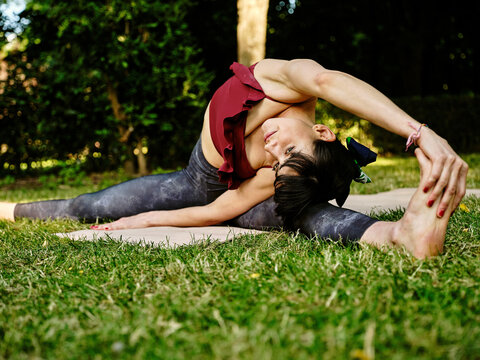 Young slim female in sportswear practicing Parivrtta Upavistha Konasana yoga asana on green grass in summer park
