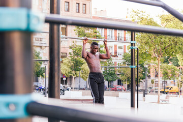 Focused African American male athlete doing pull ups on crossbar during intense training on sports ground in city