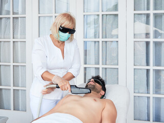 Professional woman in dark glasses and mask doing laser hair removal on chest to bearded man in salon