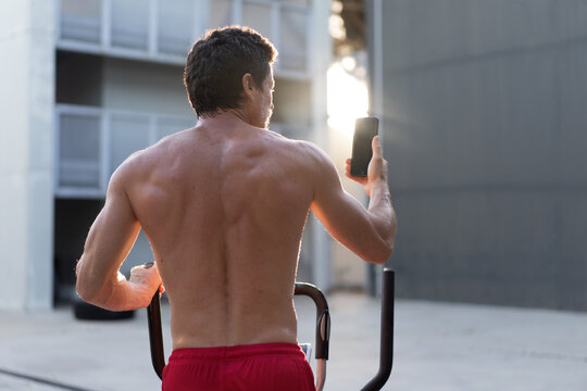 Back view of athletic male with naked torso doing exercises on stepper and taking selfie on smartphone during workout in yard