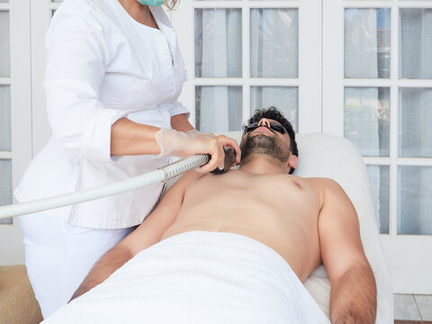 Cropped faceless master using laser apparatus on male face removing beard face during procedure in salon