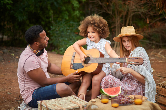 Cute curly haired little girl having fun and playing guitar during picnic with multiracial parents in summer day in nature