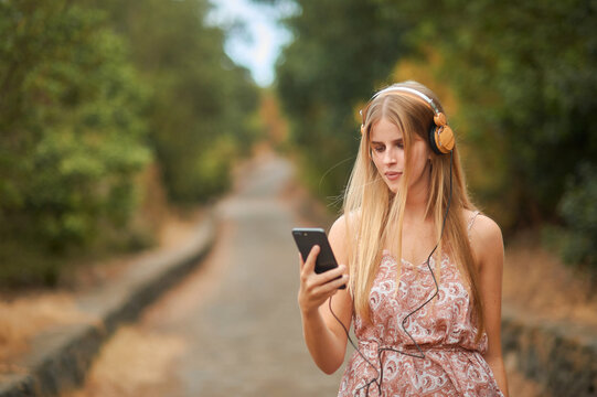 Optimistic young female in stylish summer dress and headphones enjoying music and browsing smartphone while walking on alley in park