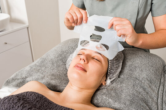Crop unrecognizable beautician removing moisturizing cloth mask from face of relaxed female client during professional treatment in modern clinic
