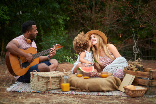 Happy young mixed race parents with cute curly haired daughter playing guitar and laughing while chilling together during picnic in summer day in green forest