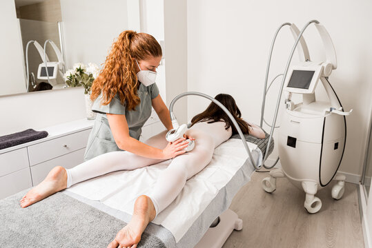 Masseuse in protective mask doing anti cellulite lipo massage to female client dressed in white suit during body care procedure in salon of apparatus cosmetology