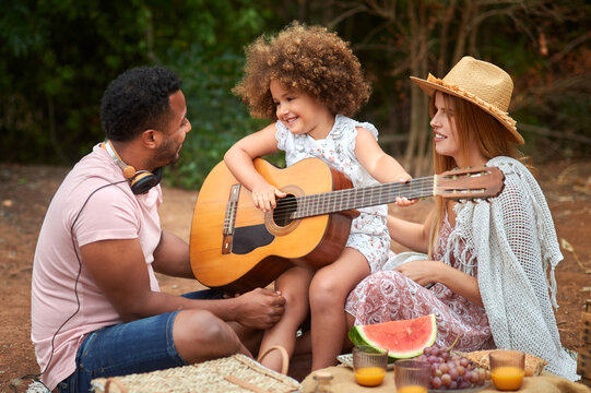 Cute curly haired little girl having fun and playing guitar during picnic with multiracial parents in summer day in nature