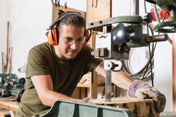 Focused male carpenter in protective ear muffs and goggles cutting piece of wood with band saw in shabby workshop