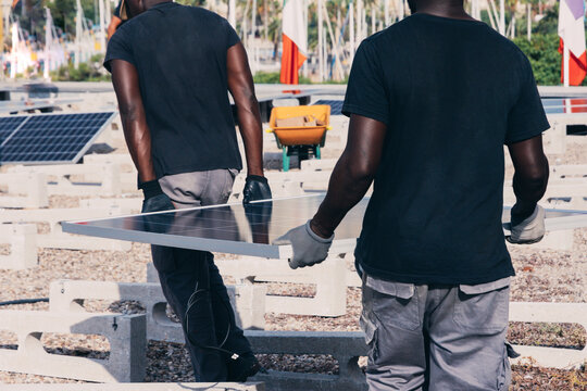 Back view of crop African American workers walking with solar panel on sunny day in industrial area
