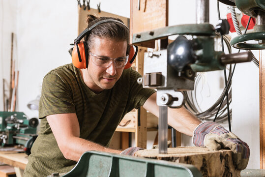 Focused male carpenter in protective ear muffs and goggles cutting piece of wood with band saw in shabby workshop
