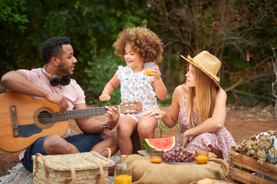 Happy young mixed race parents with cute curly haired daughter playing guitar and laughing while chilling together during picnic in summer day in green forest