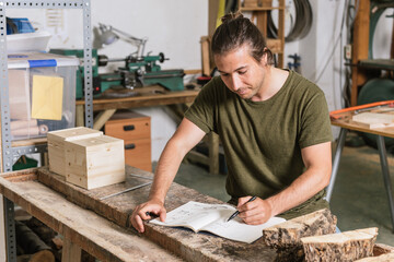 Concentrated male carpenter standing at workbench and taking notes in notebook while working in garage