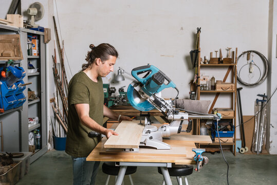 Side view of male carpenter cutting wooden plank with miter saw while working in bright workshop