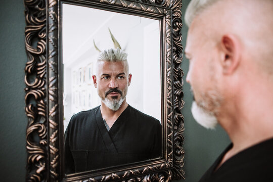 Confident brutal middle aged bearded male with gray hair in black clothes  looking at reflection in framed mirror