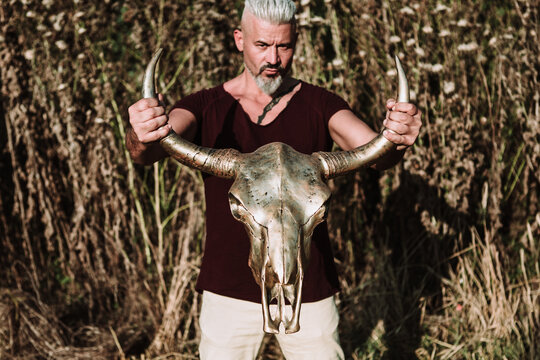 Serious muscular bearded gray haired male with tattoo holding horn animal skull while standing against tall grass and blue sky in nature looking at camera
