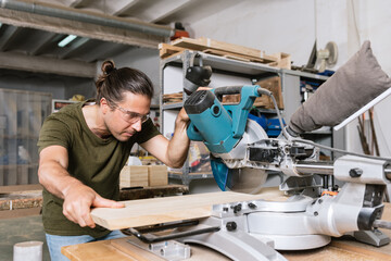 Male carpenter in protective goggles cutting wooden plank with miter saw while working in bright workshop