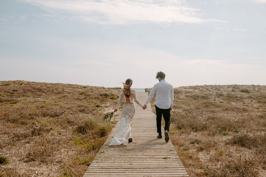 Full body back view of unrecognizable young newlywed couple in stylish clothes with bouquet holding hands and walking on wooden pathway towards sea