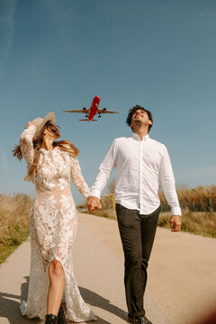 Happy young man in stylish outfit holding hand of woman in white wedding dress while strolling together on country road against blue sky with flying airplane