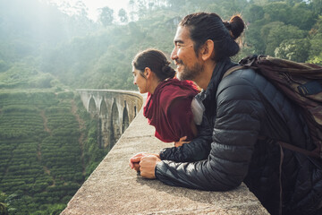 Side view of cheerful ethnic man and woman in activewear with backpacks standing contemplating views on aged stone Nine Arch Bridge among green hills in Sri Lanka