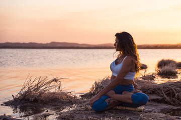 Full length serene young female in sportswear sitting in Revolved Lotus Pose while meditating during yoga practice on lake shore during sunset
