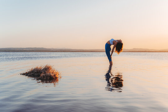 Full length side view of young flexible female in sportswear practicing yoga and doing Standing Backward pose with eyes closed in calm lake water during sunset