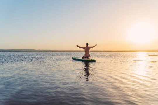 Full body back view of unrecognizable man doing yoga variation asana with open arms while floating on paddle board in calm lake water during sunset