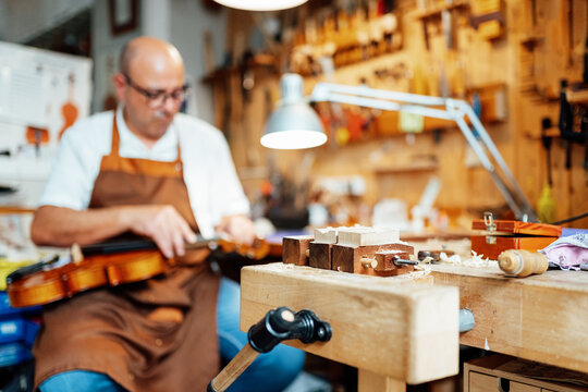 Selective focus of detail of wooden luthier workbench in workshop with blurred male artisan repairing violin