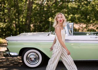 Side view of charming young blonde female in stylish outfit looking away while walking near old fashioned car in summer day