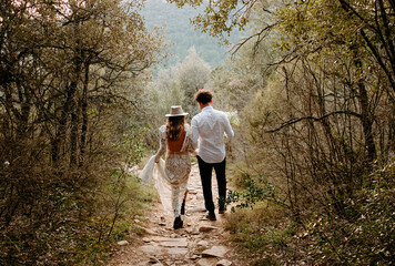 Back view of unrecognizable young bride and groom in stylish wedding outfits walking together on narrow stony path in mountainous forest in Morro de Labella in Spain