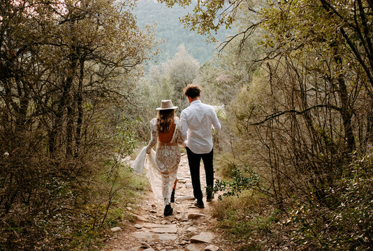 Back view of unrecognizable young bride and groom in stylish wedding outfits walking together on narrow stony path in mountainous forest in Morro de Labella in Spain