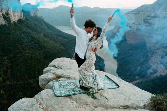 Full body side view of happy young newly married couple in elegant outfits holding colorful smoke bombs and kissing while celebrating marriage on top of rock in Morro de labella in Spain