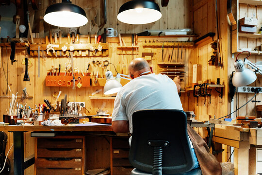 Back view of unrecognizable male luthier master sitting on chair and catching tool from wall while making instruments in workshop