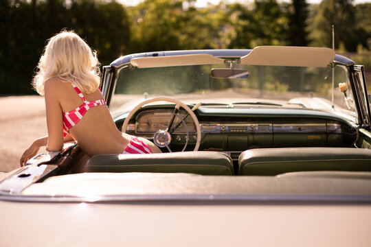 Back view of anonymous woman in striped swimwear peeking out from retro vehicle on sunny summer day in countryside