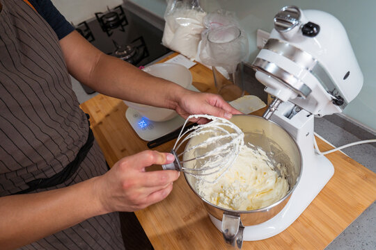 Faceless home baker removing whisk off stand mixer with bowl of whipped buttercream on counter