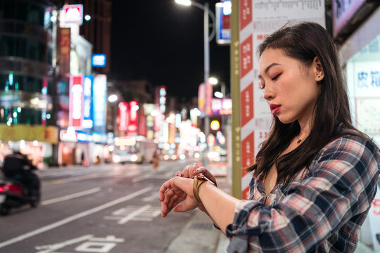 Side view of young Asian female in casual outfit checking time on wristwatch while waiting for bus on illuminated city street in evening time