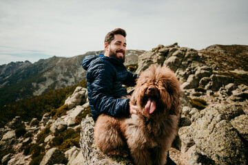 Cheerful bearded man sitting on stone near cute Labradoodle against overcast sky during trip in Puerto de la Morcuera mountains in Spain looking at camera