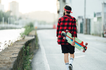 Back view of male skater in checkered shirt and with longboard walking along asphalt road while relaxing during summer weekend in city