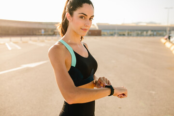 Young fit female looking at camera in sportswear checking sports results on activity tracker while resting after running on street