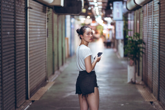 Full body young Asian female in casual clothes with mobile phone in hand walking on narrow pedestrian underground pathway with closed shops in city