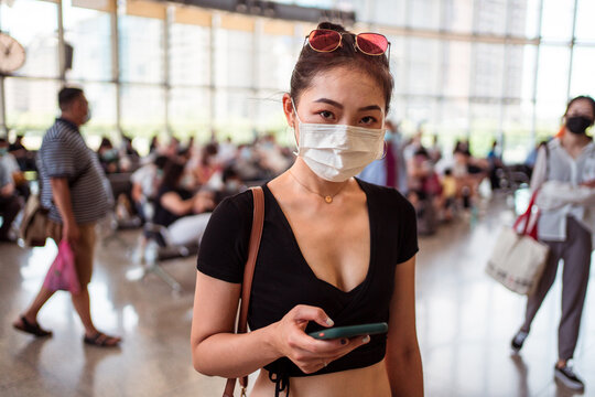 Ethnic female in protective mask standing in departure lounge of contemporary airport and surfing Internet on cellphone looking at camera