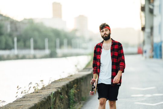 Full body of male skater in plaid shirt and bandana standing with longboard on empty road and looking away