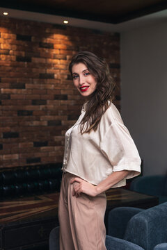 Side view of charming confident young female with red lips and long wavy hair dressed in stylish blouse and pants looking at camera while standing near chair in modern room with loft interior