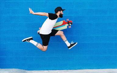 Side view of talented male skater with longboard in moment of jumping on background of blue wall in city