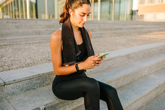 Young slim female in sportswear with earbuds and fitness tracker using mobile phone while standing on street after training