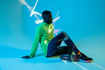 Full body side view of strong young African American male athlete in stylish colorful tracksuit and sneakers sitting on floor in blue studio with neon lights