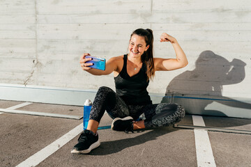 Cheerful female athlete in sportswear sitting on street after training and taking photo on selfie camera
