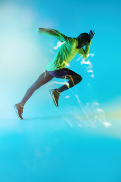 Full body side view of young African American male runner in colorful tracksuit sprinting from start position in studio with bright neon illumination