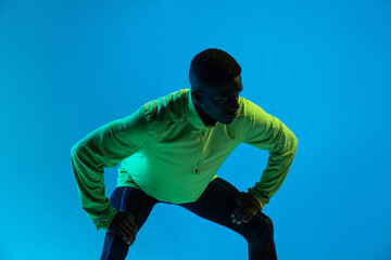 Strong young African American male athlete in stylish colorful tracksuit and sneakers standing in blue studio